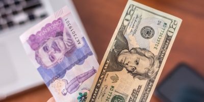 Hand holding dollar bill and colombian peso with smartphone and laptop in background