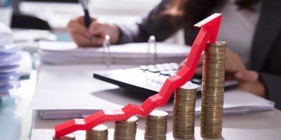 Increasing Golden Coins With Red Arrow Showing Upward Direction In Front Of Businessperson Calculating Bill
