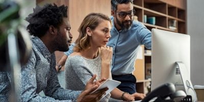 Focused businesspeople discussing project while looking at computer screen. Mature manager working with his creative team in a modern office. Multiethnic casual teamwork at work.