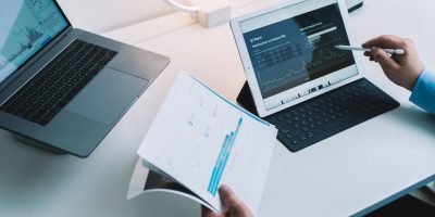 From above of crop faceless male financier read documents while working on tablet and laptop while checking change of stock market in office