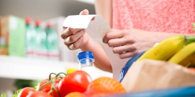 Unrecognizable woman checking a long supermarket receipt with grocery on foreground.